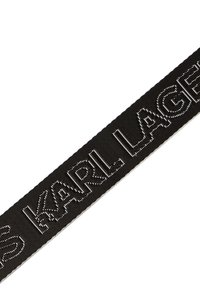 Cinta de tela negra con letras plateadas en relieve que dicen "KARL LAGERFELD". Superficie texturizada con un brillo suave. Bordes acabados en blanco.