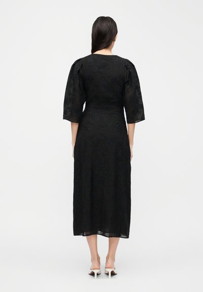 Γυναίκα με μακριά σκούρα μαλλιά, φορώντας μαύρο φόρεμα μεσαίου μήκους και φουσκωτά μανίκια τριών τετάρτων, όρθια με την πλάτη, εμφανείς λευκές σανδάλες με τακούνι.