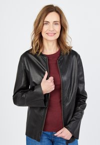 Mustapintainen nahkatakki, jossa on pyöreä kaulus ja vetoketjukiinnitys. Takissa on pitkät hihat ja se on rento malli, joka on päällä viininpunaisen topin päällä.