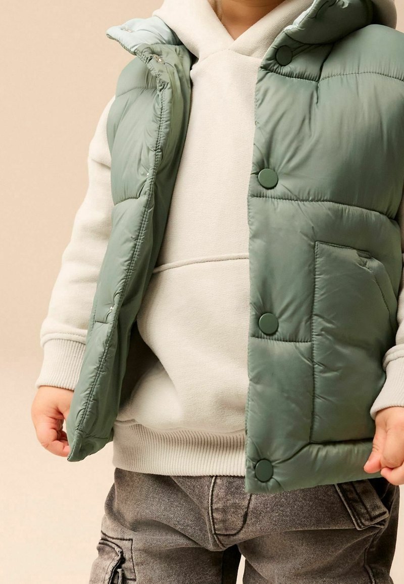 Grüne gepolsterte Weste mit Druckknöpfen, die über einem hellgrauen Hoodie getragen wird. Graue Denim-Hosen mit Cargo-Taschen. Insgesamt glatte Textur.