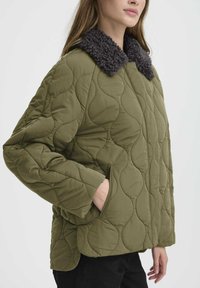 Giacca trapuntata verde oliva con un colletto in pelliccia grigia scura texturizzata. Presenta tasche laterali e un design a vestibilità rilassata.