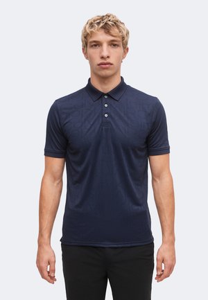 Giovane uomo con capelli biondi corti e ricci, che indossa una polo blu scuro a maniche corte e pantaloni neri, rivolto in avanti con espressione neutra.