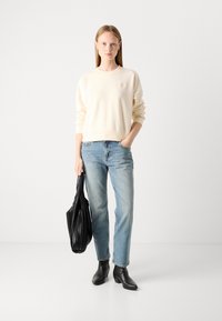 Sweatshirt crème avec des poignets et un ourlet côtelés, jeans bleu clair, bottines noires à la cheville et un sac fourre-tout texturé noir, se tenant sur un fond uni.