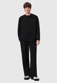 Young man wearing a black long-sleeve sweatshirt and black wide-leg pants with white and black sneakers, standing against a plain background.