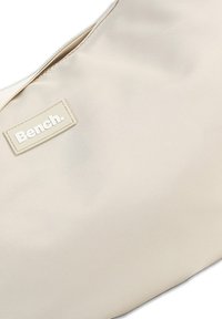 Beige Nylon Schultertasche mit glatter Textur, ausgestattet mit einem kleinen rechteckigen Logo-Patch, auf dem "Bench." steht, und einem Reißverschluss.