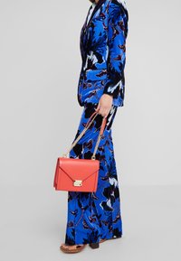 Blå velour dragt med blomsterprint, parret med en rød lædertaske med guldfarvede metaller og kædestrop.