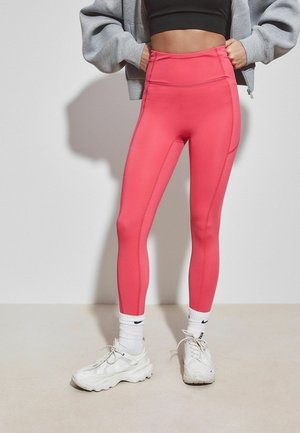 Personne portant des leggings taille haute rose vif, des baskets blanches Nike, des chaussettes blanches, un soutien-gorge de sport noir et une veste grise zippée, debout contre un mur uni.