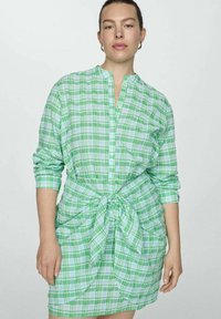 Abito camicia a quadri verde e blu con cintura da annodare, maniche lunghe e chiusura frontale con bottoni. Tessuto leggero con una consistenza morbida.
