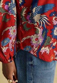 Rote Bluse mit floralem Muster und blauen sowie pinken Akzenten, aus leichtem Stoff mit gerafften Ärmeln, kombiniert mit Jeans. Das florale Muster ist deutlich sichtbar.