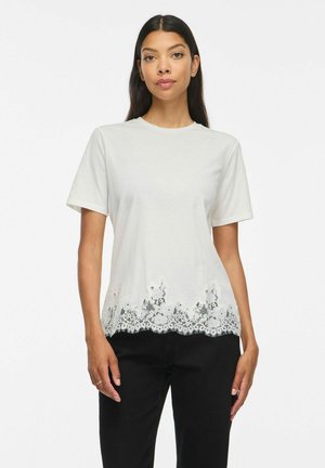 Femme aux longs cheveux foncés portant un tee-shirt blanc à manches courtes avec un détail en dentelle en bas et un pantalon noir sur fond blanc.