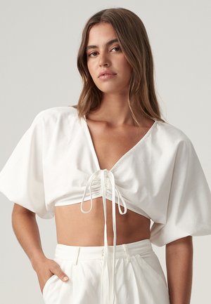 Top bianco cropped con maniche corte a sbuffo, scollatura a V e dettagli con lacci sul davanti. Realizzato in tessuto leggero, che mostra una consistenza liscia.