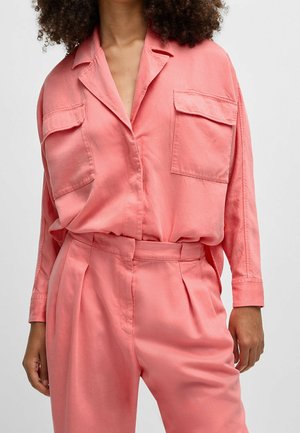 Woman wearing a loose-fitting coral pink shirt with two chest pockets and matching high-waisted pants against a plain background.