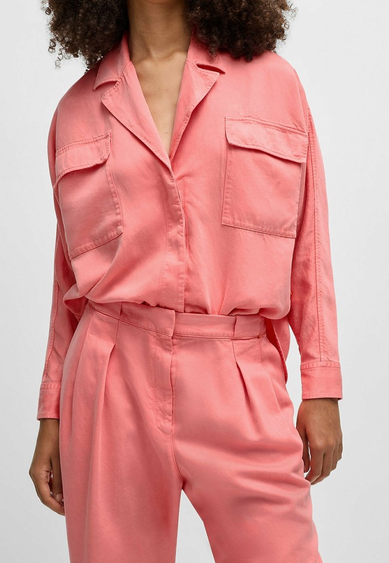 Femme portant une chemise ample rose corail avec deux poches poitrine et un pantalon taille haute assorti sur un fond uni.