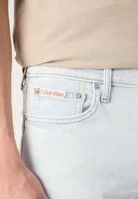 Svijetloplave traperice glatke teksture, s crvenom oznakom "Calvin Klein", srebrnim hardverom i klasičnim dizajnom s pet džepova.