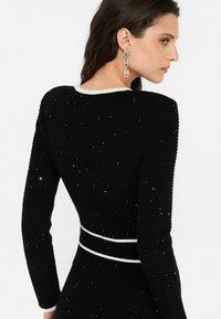 Abito nero strutturato con maniche lunghe, con rifiniture bianche all'escursione e in vita, adornato da abbellimenti sparsi.