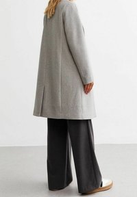 Long manteau gris à coupe droite, avec une fente au dos, porté sur un pantalon large foncé et des chaussures de couleur claire. Texture lisse.