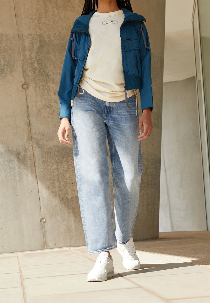 Modrá zipová bunda přes béžové tričko, doplněná o světle modré široké džíny a bílé tenisky. Hladké textury tkanin a volný střih.