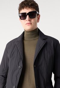 Chaqueta ligera negra con un acabado texturizado, con un cuello de solapa y dos botones frontales, llevada sobre un suéter de cuello alto verde oscuro. Gafas de sol cuadradas negras.