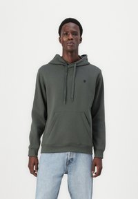 Sweat à capuche vert foncé en tissu doux, doté d'une poche kangourou, d'une capuche avec cordon réglable et d'un petit logo sur la poitrine.