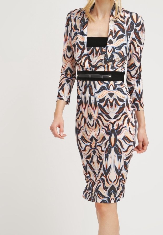 Femme portant une robe longueur genoux avec un motif abstrait multicolore et une ceinture noire, debout devant un fond clair uni.