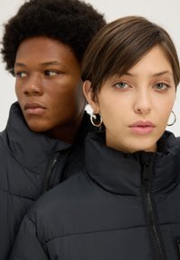 Close-up of a man and woman wearing black puffer jackets, woman with short hair and hoop earrings facing forward, man looking sideways.