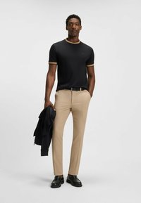 Czarna koszula z krótkim rękawem z beżowymi akcentami, beżowe spodnie i czarne buty. Model trzyma czarną kurtkę na neutralnym tle.