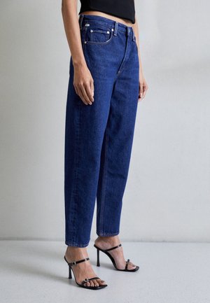 Personne portant un jean droit bleu foncé taille haute et des sandales noires à talons hauts ouvertes aux orteils, debout contre un mur uni.