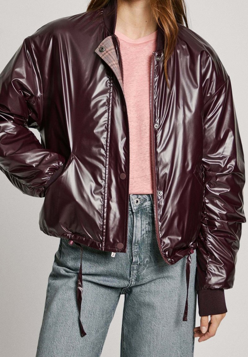 Femme portant une veste zippée brillante de couleur bordeaux foncé sur un t-shirt rose clair et un jean taille haute bleu clair, debout avec une main dans la poche.