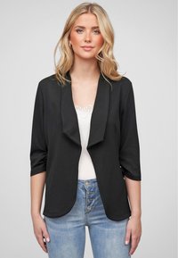 Svart, teksturert blazer med sjalkrage, trekvart lange ermer og åpen front, kombinert med en hvit topp og høyreiste lyseblå jeans.