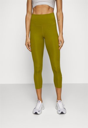 Personne portant des leggings courts taille haute couleur vert olive, un soutien-gorge de sport blanc et des baskets blanches, debout devant un fond blanc uni.
