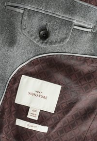 Grijze stoffen binnenkant van een colbert met een donker bordeaux rode voering met een gegraveerd geometrisch patroon, labels voor maat en merk.