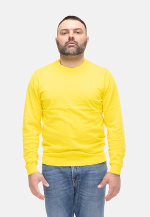 Uomo con capelli corti e barba che indossa una maglietta gialla a maniche lunghe e jeans blu, in piedi davanti a uno sfondo bianco semplice.