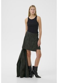 Schwarzes Tanktop, grüne strukturierte Rock mit vertikalen silbernen Linien, asymmetrischer Schnitt, kombiniert mit knielangen schwarzen spitzen Stiefeln.