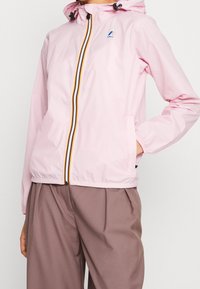 Lyserød hættejakke med stripet lynlås og logo på brystet, båret med brune plisserede bukser, hånd i lommen, på en person.