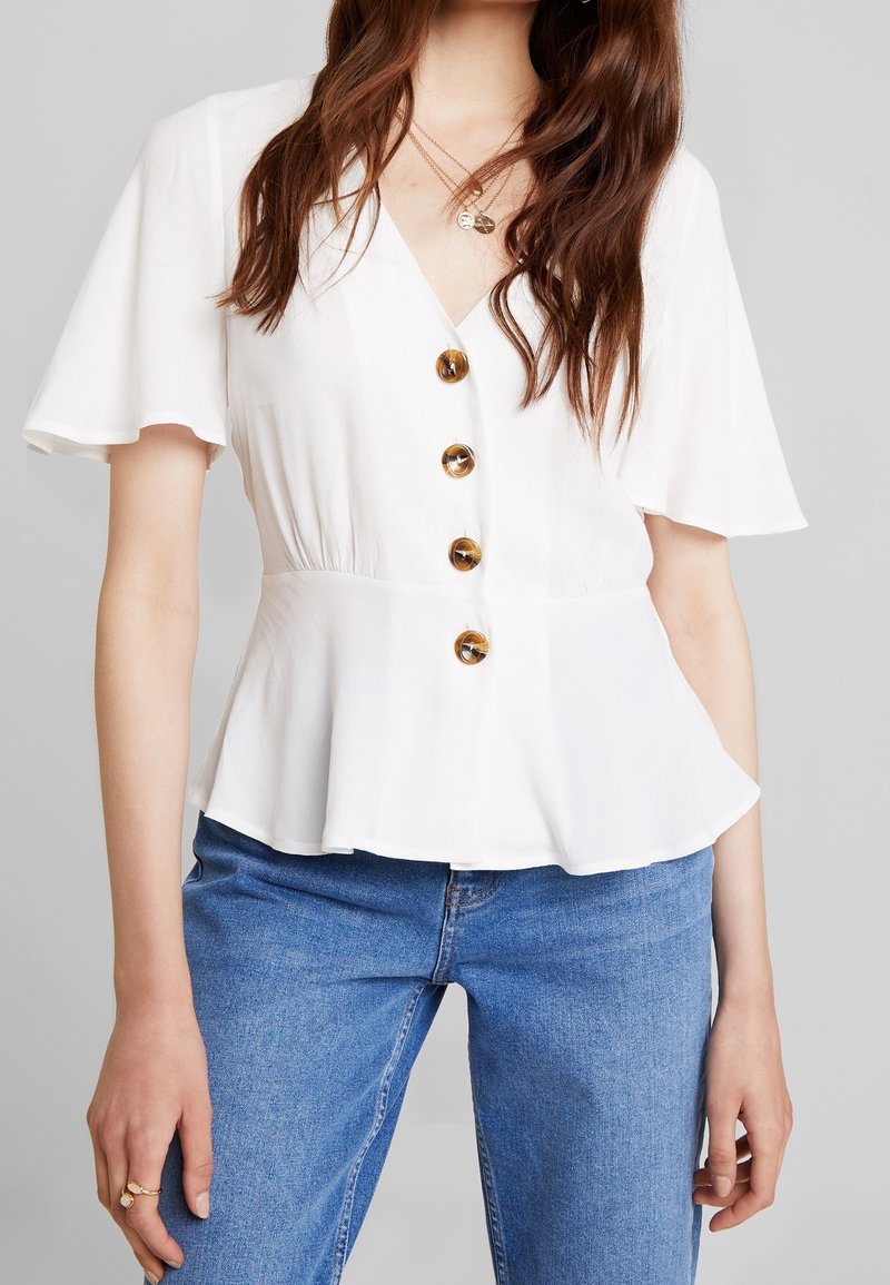 Femme portant un chemisier blanc à manches courtes avec quatre gros boutons marron et un jean bleu, montrant le torse et les bras.