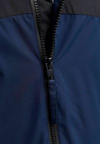 Námornícka bunda na zips s čiernou hornou časťou, s viditeľným plastovým zipsom na prednej strane a hladkou, matnou textúrou.