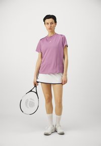 Šviesiai purpurinė sportinė marškinėliai su trumpomis rankovėmis, derinami su balta sijonu, turinčiu tamsiai mėlyną akcentą. Modelis laiko juodą teniso raketę.