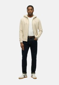 Felpa beige con zip sopra una maglietta bianca, abbinata a jeans scuri e sneaker bianche con dettagli neri. Tessuto di cotone morbido e vestibilità casual.
