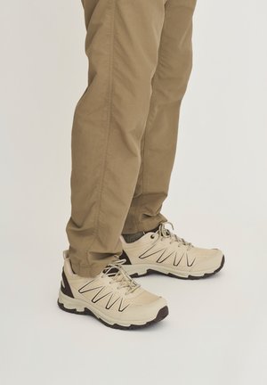 Beige hiking shoes with black accents worn with brown pants and dark gray socks on a light background.