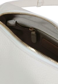 Witte leren tas met textuur en een gouden ritssluiting die deels openstaat, waarbij de binnenvoering van stof en een binnenzak met ritssluiting en leren trekker zichtbaar zijn.