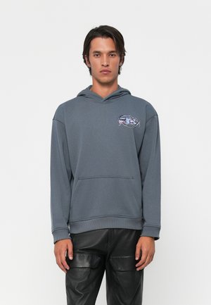 Young man wearing a gray hooded sweatshirt with a small abstract graphic on chest and black leather pants against a white background.