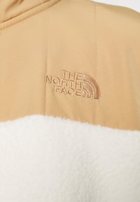Kahvin ja valkoisen eristetyn takin alaosa on pehmeää fleeceä ja yläosa sileää kangasta, jossa on kahvinvärinen "The North Face" -logo brodeerattuna.