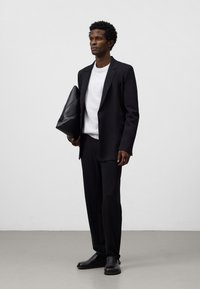 Giacca nera sopra un maglione grigio chiaro e pantaloni abbinati. Una pochette nera di tessuto è tenuta nella mano sinistra. Scarpe nere completano il look.