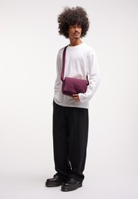 Borsa a tracolla viola scuro con una texture liscia, forma rettangolare e tracolla regolabile. Indossata con una camicia bianca a maniche lunghe e pantaloni neri.