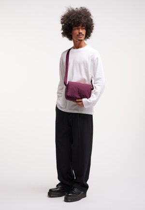 Borsa a tracolla viola scuro con una texture liscia, forma rettangolare e tracolla regolabile. Indossata con una camicia bianca a maniche lunghe e pantaloni neri.