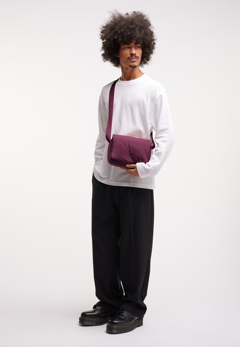 Borsa a tracolla viola scuro con una texture liscia, forma rettangolare e tracolla regolabile. Indossata con una camicia bianca a maniche lunghe e pantaloni neri.