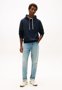 Sudadera con capucha azul marino con cordones blancos, bolsillo canguro, combinada con vaqueros azul claro y zapatillas blancas con suelas color canela.