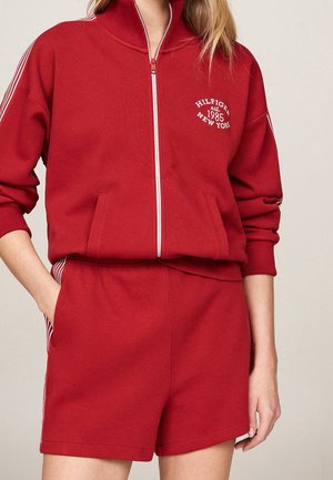 Kobieta ubrana w czerwoną kurtkę zapinaną na zamek z napisem „Hilfiger est. 1985 New York” oraz pasujące czerwone szorty z białymi paskami po bokach.