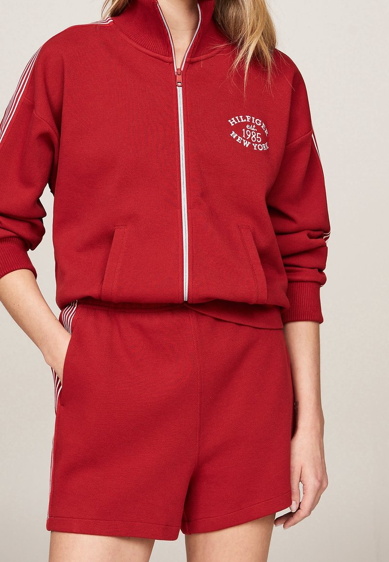 Femme portant une veste rouge zippée avec le texte « Hilfiger est. 1985 New York » et un short rouge assorti avec des bandes blanches sur les côtés.