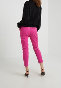 Woman stands facing away wearing bright pink pants, black long-sleeve top, and black open-back high-heeled shoes on white floor.